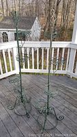 Two matching green metal indoor/outdoor plant stands on a wooden porch deck, each with three circular holders and decorative scrollwork legs and tops.