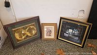 Photo of three framed wall decorations displayed side by side against a wall, showing a glass-encased world map, small floral print, and floral clay artwork.