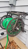 Wall-mounted metal hose reel with decorative side cut-outs and green garden hose wound on it, mounted on exterior house siding.