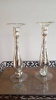 Pair of tall glass Pier One candlestick holders shown standing on a table against a plain wall