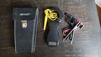 Amprobe electrical meter with yellow test lead, additional test probes, and black stitched carrying case with metal clasp on a wooden surface.