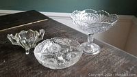 Three clear glass pieces on a dark surface including a covered oval dish, pedestal compote bowl, and small footed bowl featuring intricate pressed glass patterns.