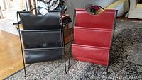 Photo showing a pair of leatherette magazine holders; one black and one red, on a patterned carpet floor.