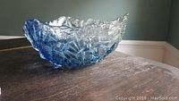 Side angle of a handmade glass bowl showing clear and blue textured, geometric design with irregular ruffled edges, placed on a dark wooden surface.