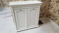 White wooden bathroom cabinet with two front panel doors and small knobs, standing on a tiled floor next to floral wallpapered wall.
