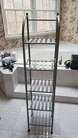 Photo showing the full metal towel rack standing on tiled floor in front of a bathtub with window background. Four shelves with slatted metal design, silver finish.