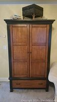 Front view of the tall armoire showing two paneled doors and drawer at the bottom with black frame and brown wood panels.