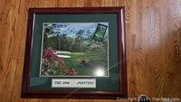 Framed print showing a colorful golf course scene from the 2006 Masters with two small attached items in top corners.