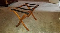 Wooden luggage holder with four black straps, curved legs, and natural wood finish set on carpeted floor.