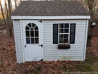 Front view of white vinyl-sided shed with arched window in door, black shutters and planter box under window