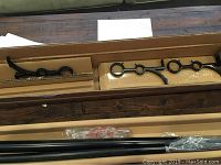 Two black metal curtain rods with mounting brackets laid in cardboard boxes on wooden floor, with plastic wrapping on hardware parts.