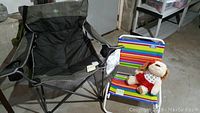 Wide view showing the REI folding camp chair in gray and black, smaller beach chair with colorful stripes, and plush dog toy seated on the smaller chair, all set on a concrete basement floor.