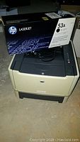 HP LaserJet P2015 printer with the sealed HP 53A toner cartridge box placed on top. Printer is beige with dark gray top and control panel with several buttons.
