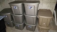 Eight Sterilite 18-gallon storage tubs with lids stacked against a wall, two additional similar sized storage bins nearby, including one black and one beige bin.