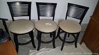 Set of three black swivel bar height chairs with padded beige seats and horizontal slatted backrests. Chairs show visible wear including stains and scuffs.