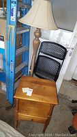 Floor lamp and wooden nightstand positioned next to a blue ladder and black chairs in a garage environment.