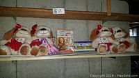 Four Martha Stewart plush girl dogs with red bows and skirts, a pink Ty Beanie Baby unicorn, and two textured painting kits on shelf in concrete room.