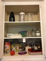 Three-shelf cabinet filled with kitchen items including Veggetti Pro box, oven dishes, glass jars, thermos, pitcher, and other kitchenware.