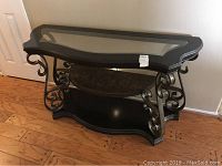 Front view of a hall or sofa table with glass top, metal sides with scrollwork, and wooden bottom shelf