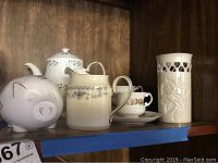 Shelf with white piggy bank, teapot, creamer, cups with saucers, and tall vase.