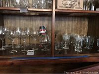 Wide shot of all glasses arranged on two wood shelves showing wine, cognac, water with leaf design, dessert, and martini glasses with price tag visible.