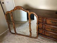 Image of a large decorative wood-framed mirror with scalloped edges and an ornate top carved design, placed on carpeted floor against a wall.