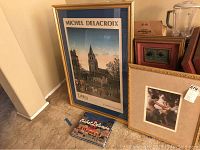 Framed Michel Delacroix print 'Lublin' 39x27 inches, displaying a cityscape scene with a tall tower, horse-drawn carriage and figures, next to a Michel Delacroix art book and other framed artworks.