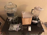 KitchenAid blender, glass beverage dispenser with stand, wooden box with clear plastic buffet plates, metal wine glass holders and table cloth holders on a black surface.