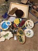 Overview of handpainted ceramic serving plates featuring fruit, olive, and flower motifs, wooden basket trays, a clear glass bowl, and metal utensils.