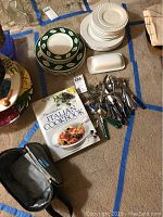 Overview image showing stacked plates grouped by design and pattern, cutlery set, butter dish, and Italian cookbook on carpet.