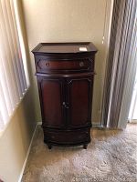 Full front view of a wooden jewelry cabinet with multiple drawers and storage doors against a wall.