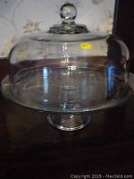 Clear glass cake stand with dome cover on a wooden surface, showing overall shape and condition.