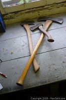 Three vintage axes lying on wooden surface near window; wooden handles with metal blades, one axe is double-headed.