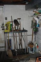 Wide view of assortment of long-handled vintage garden and home tools leaning against white paneled wall including shovels, rakes, hoes, broom, and axe.