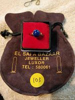 The ring displayed in its case atop the brown drawstring bag from El Safa Bazaar Luxor with contact number printed in gold.