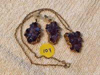 Amethyst earrings and matching pendant laid on a textured fabric with chain loosely arranged. Yellow sticker with number 107 visible.