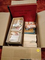 Top-down view of a cardboard box containing a red hardcover postage stamp album and two smaller boxes filled with envelopes and loose stamps.