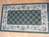 Full view of the green and white small rectangular carpet with a patterned center and leaf border on wooden floor.