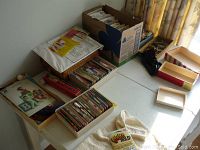 Overview of white tray and cardboard box filled with vintage sewing patterns and baskets with sewing accessories on table.