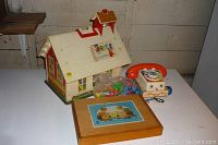 Vintage Fisher Price Toy School house, rotary dial pull toy phone, a bag of small toy parts, and wooden puzzle box placed on a white table.