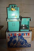 Vintage teal Easy-Bake Oven approximately 14 inches tall with clock decal peeling on top and Easy-Bake Oven decal on front. Shows vintage wear.