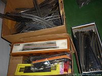 Box containing various vintage toy train tracks, including curved and straight pieces, and a boxed Tyco train car.