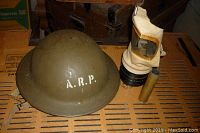 WWII A.R.P. helmet, gas mask, and brass round casing arranged on a surface. The helmet is olive green with 'A.R.P.' lettering on front. Gas mask has beige fabric hood with clear window and black canister. Brass casing standing upright.