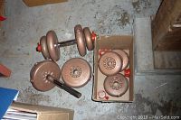 Top-down view showing several bronze-colored metal weight plates and two weight bars, one assembled with plates, one loose, and a cardboard box containing extra plates.