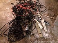 Several black and orange extension cords tangled together on concrete floor, some with attached power strips.
