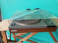 Turntable with closed dust cover on wooden base against teal wall.