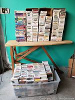 Full view of original VHS tapes in reduced boxes stacked on an ironing board and clear plastic bin.
