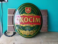 Front view of the oval Okocim beer sign on a wooden surface, showing wooden accents on each side and attached cable.
