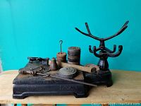 Black cast iron scale with assorted weights and a metal measuring tool on table against turquoise wall.