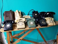 Photo showing a variety of vintage rotary dial and push button telephones arranged on a wooden table against a teal wall, including a brown car phone case phone, different colored phones in black, beige and green, with cords attached.